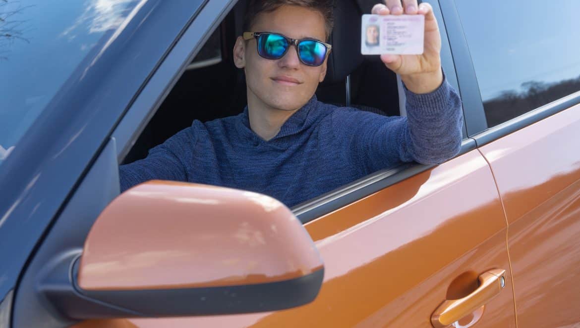 Happy teenager is showing his driving license from open car window. Driving school concept